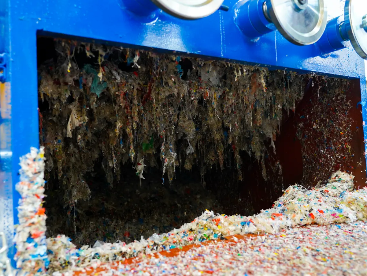 um close-up de uma máquina usada no processo de reciclagem, especificamente o que parece ser o interior de um triturador ou moedor. No interior, podemos ver fragmentos de plástico triturados, que são o resultado do processamento da máquina de pedaços maiores de resíduos plásticos em tamanhos menores e mais gerenciáveis. O processo de trituração é essencial na reciclagem de plásticos, pois prepara o material para etapas posteriores, como lavagem, separação e, eventualmente, derretimento para formar novos produtos plásticos. O plástico triturado mostrado tem cores variadas, indicando que a máquina processou uma mistura de diferentes tipos de materiais plásticos. A limpeza das lâminas e do interior da máquina é crucial para uma operação eficiente e para evitar a contaminação entre diferentes tipos de plásticos, o que pode ser crítico para a qualidade do material reciclado. A máquina parece estar em operação, com o plástico triturado saindo da máquina, pronto para passar para o próximo estágio da reciclagem.