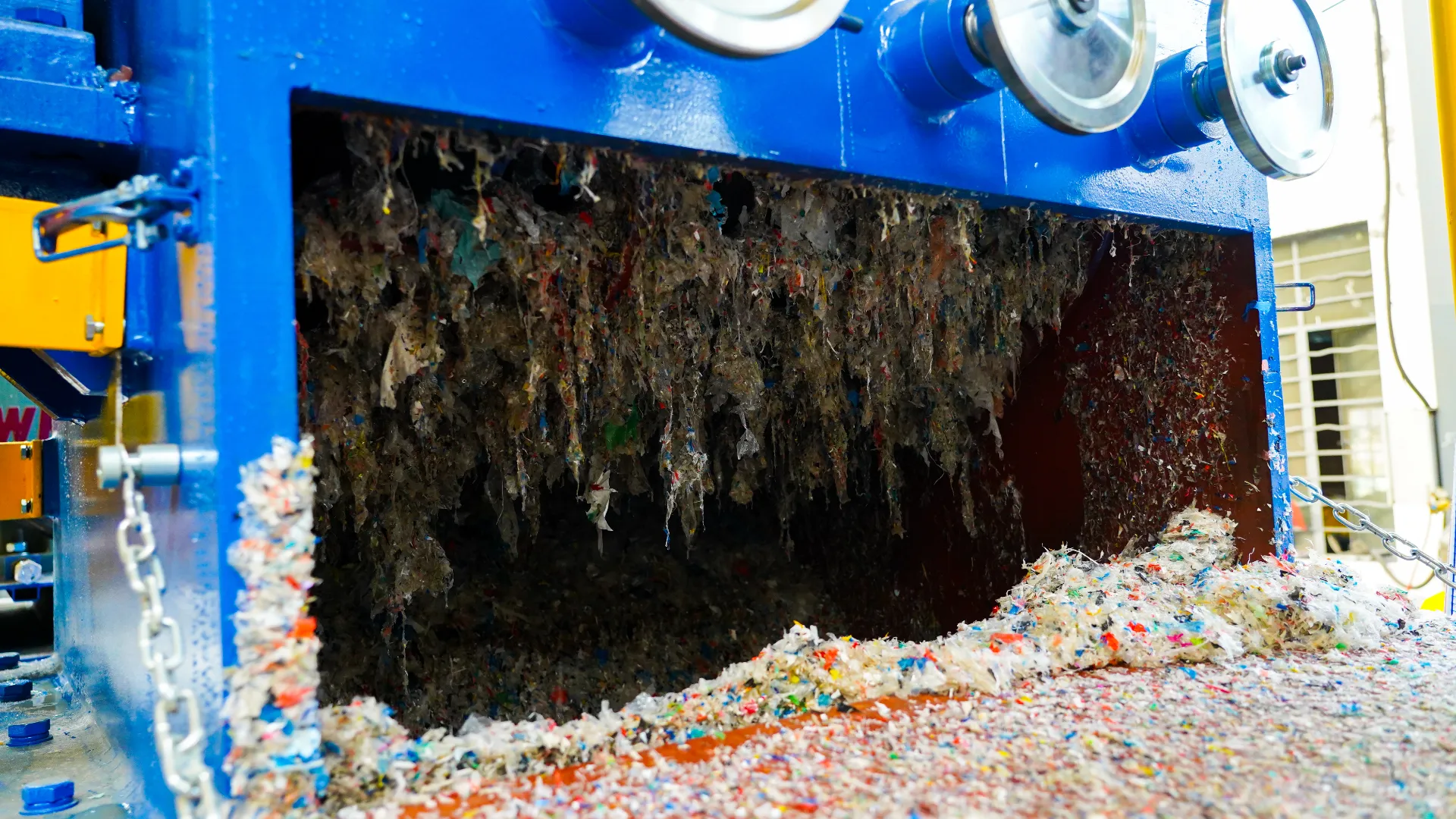 An industrial shredder in operation, processing a large amount of mixed plastic waste. The image shows shredded plastic materials hanging from the cutting mechanism inside a blue and yellow machine. Below, a pile of finely shredded plastic pieces accumulates, demonstrating the shredder's effectiveness. The equipment is designed for heavy-duty recycling processes, emphasizing durability and efficiency in handling various types of plastic waste. This shredding process is a critical step in converting plastic waste into reusable materials.