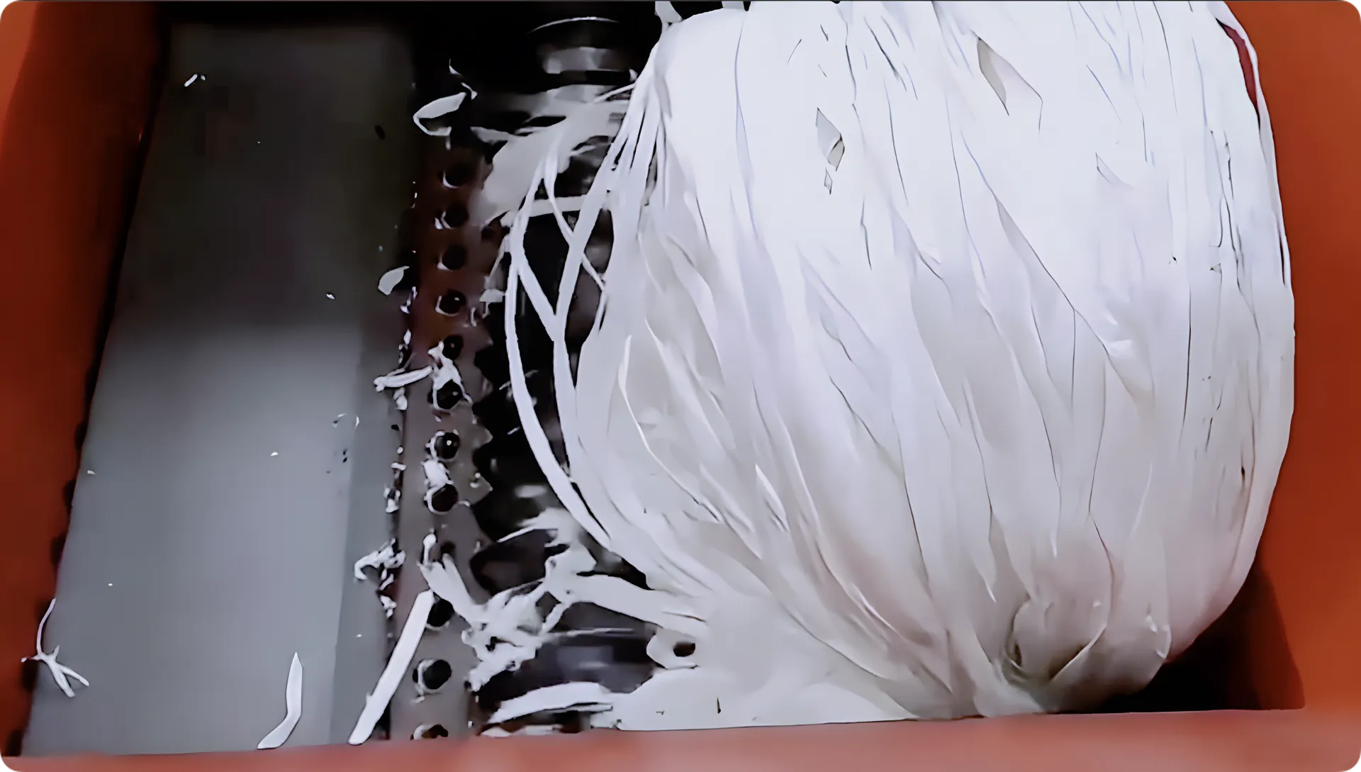 This image shows the interior of a waste film shredding machine in action. White plastic film is being fed into the machine, where it is caught by the rotating shredding mechanisms that are partially visible. These mechanisms are likely metal shafts outfitted with shredding or cutting blades, designed to break down the plastic into smaller pieces for recycling purposes. The shredding process is depicted in a dynamic state, with strips of plastic being pulled and torn by the machine, against a backdrop of a sturdy, orange housing of the shredder.