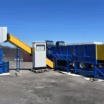 a mobile industrial shredding system for pipes, set in a barren outdoor area. The system is primarily composed of several interconnected units painted in vibrant blue and yellow, enhancing visibility. On the left, a worker in a bright orange suit and white helmet is operating the machinery, which includes a loading platform, a shredding unit, and a sorting conveyor. The clear blue sky and the lack of vegetation in the background suggest a remote or industrial location.