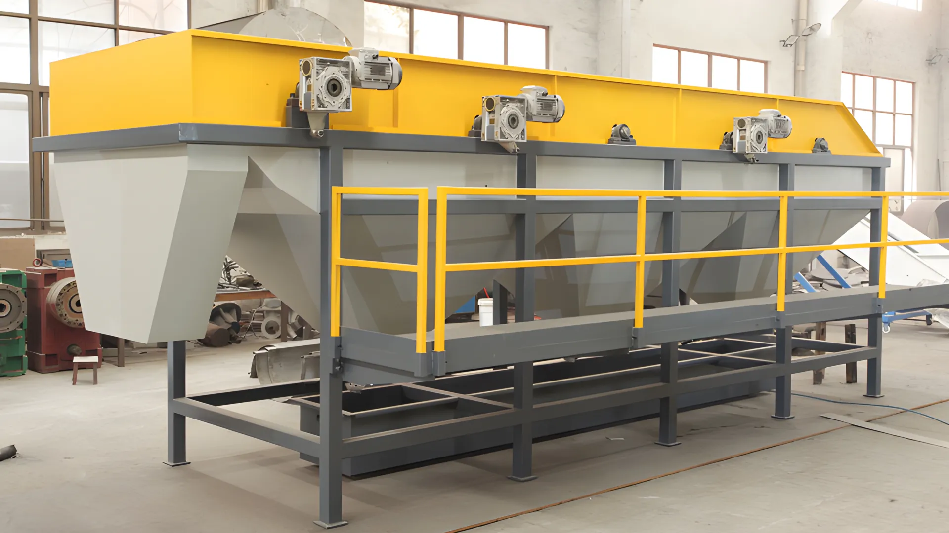 an industrial sink-float separation tank in a manufacturing facility. The tank is elongated, with a vibrant yellow top and gray structural components. Multiple electric motors mounted on top of the tank suggest its operational functionality. The structure is supported by a robust, dark gray metal frame with safety railings painted in yellow. The background of the photo indicates an industrial setting with various machinery and tools scattered around. The floor is concrete, and natural light illuminates the space through windows visible at the top.